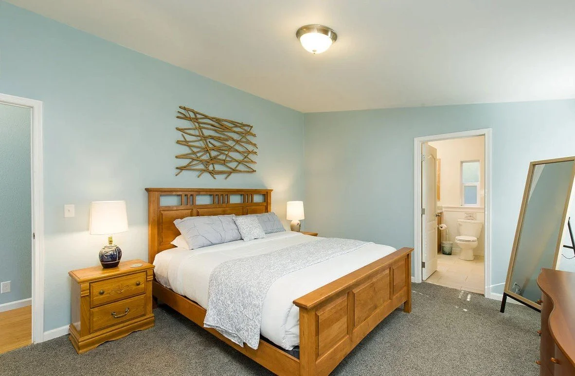 A bedroom with a wooden bed, nightstands, and lamps, and a view of a bathroom through an open door, with a mirror on the right side.