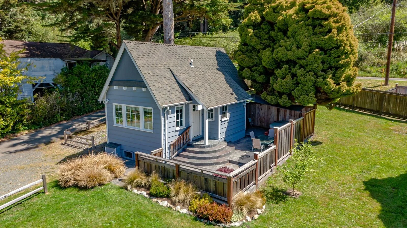 A small blue house with a gray shingled roof, front porch steps, and a fenced backyard with outdoor seating and landscaping, surrounded by trees and greenery.
