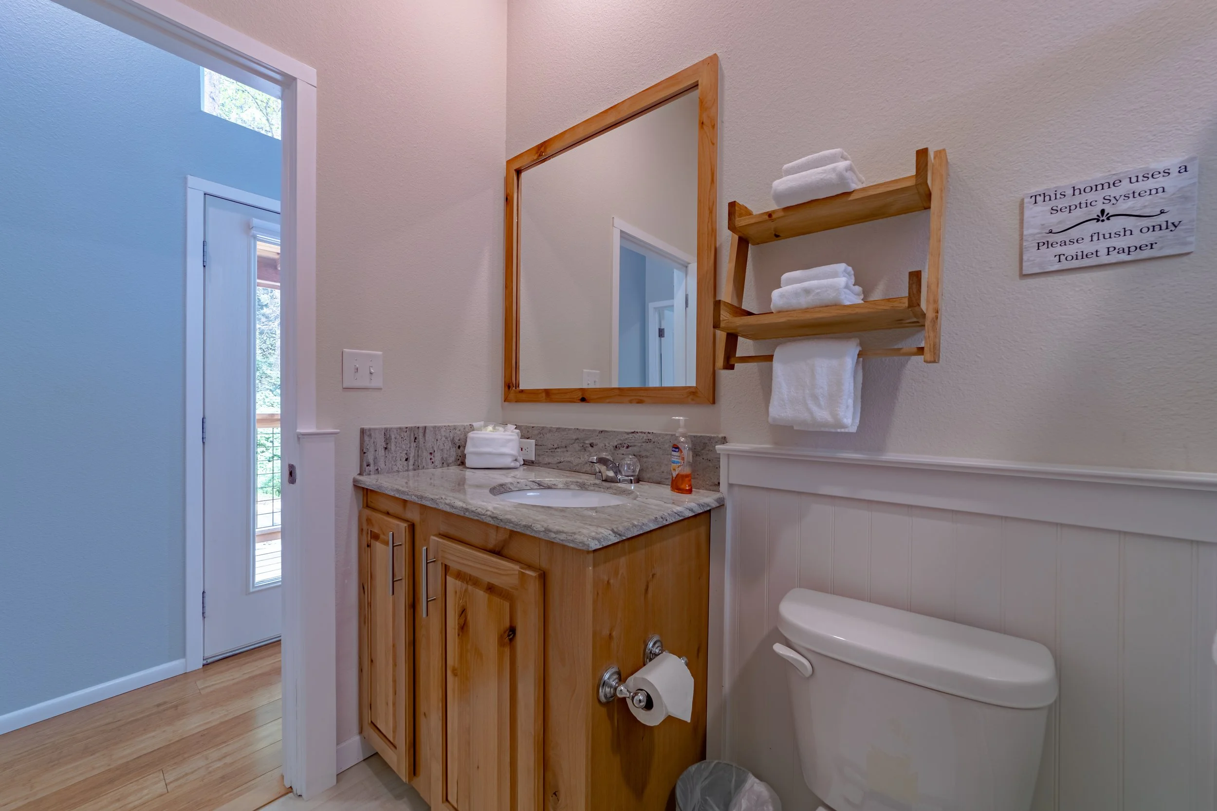 Bathroom with wooden vanity and granite countertop, mirror, toilet, wooden shelves with towels, sign about septic system, and a door leading outside.