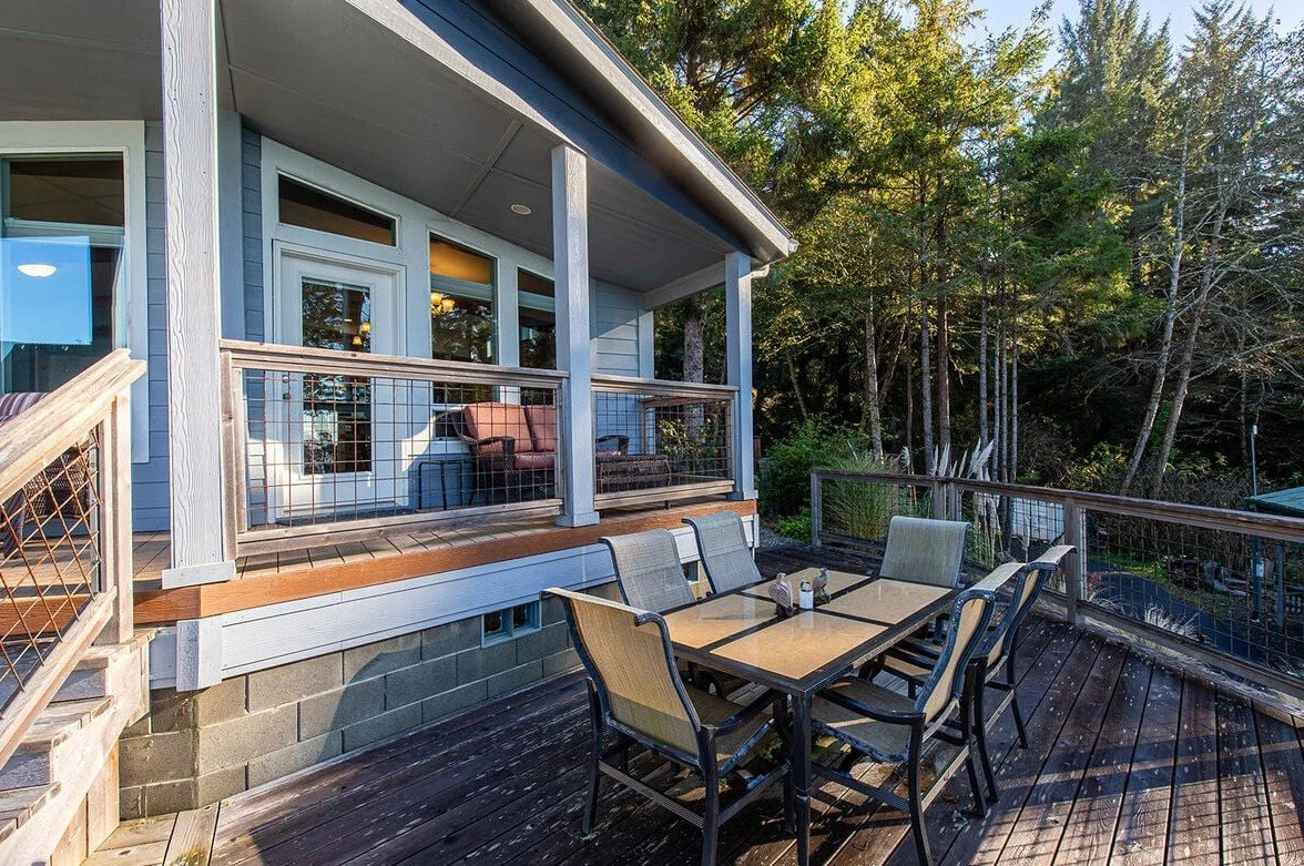 Outdoor deck with a table and six chairs, surrounded by a railing, attached to a house with a porch and seating area, overlooking trees and greenery.