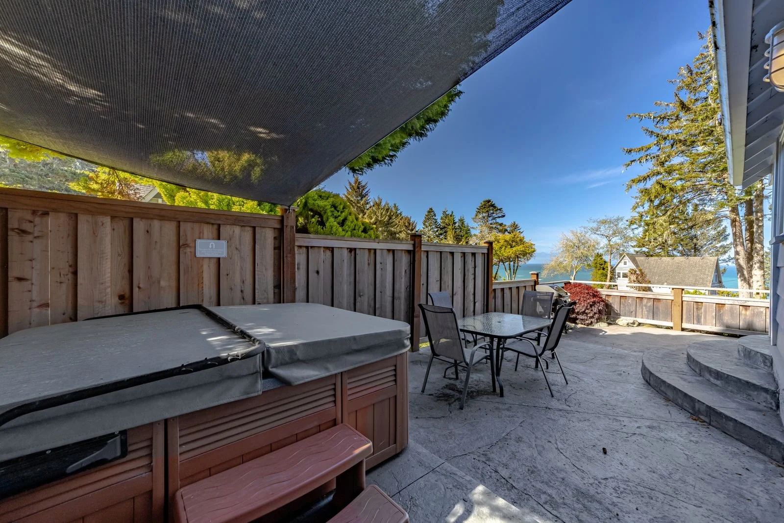 Outdoor patio area with hot tub, glass table, four chairs, wooden fencing, trees and view of water and houses in the distance.