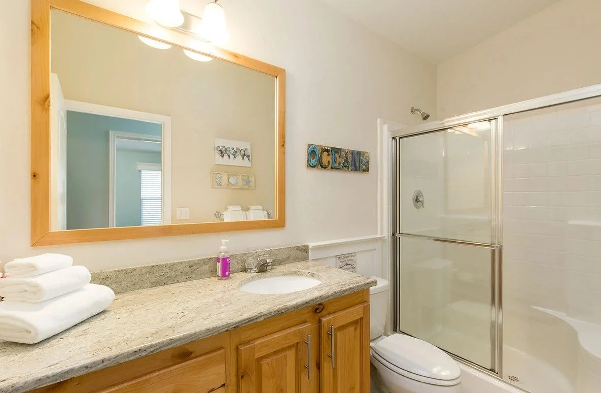 Bathroom with a wooden vanity, granite countertop, large mirror, towels, and a shower with a sliding glass door.