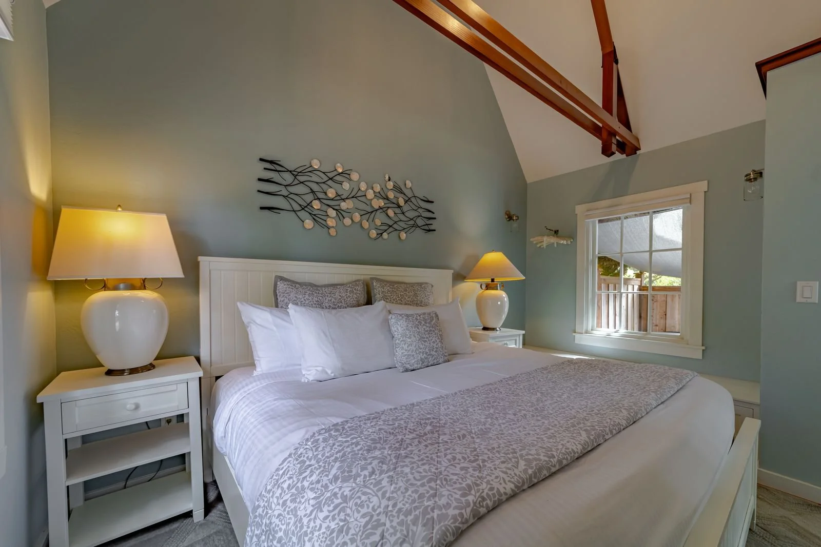 Decorative bedroom with a white bed, two nightstands with lamps, wall art, and a window with a view of a wooden fence and garden.