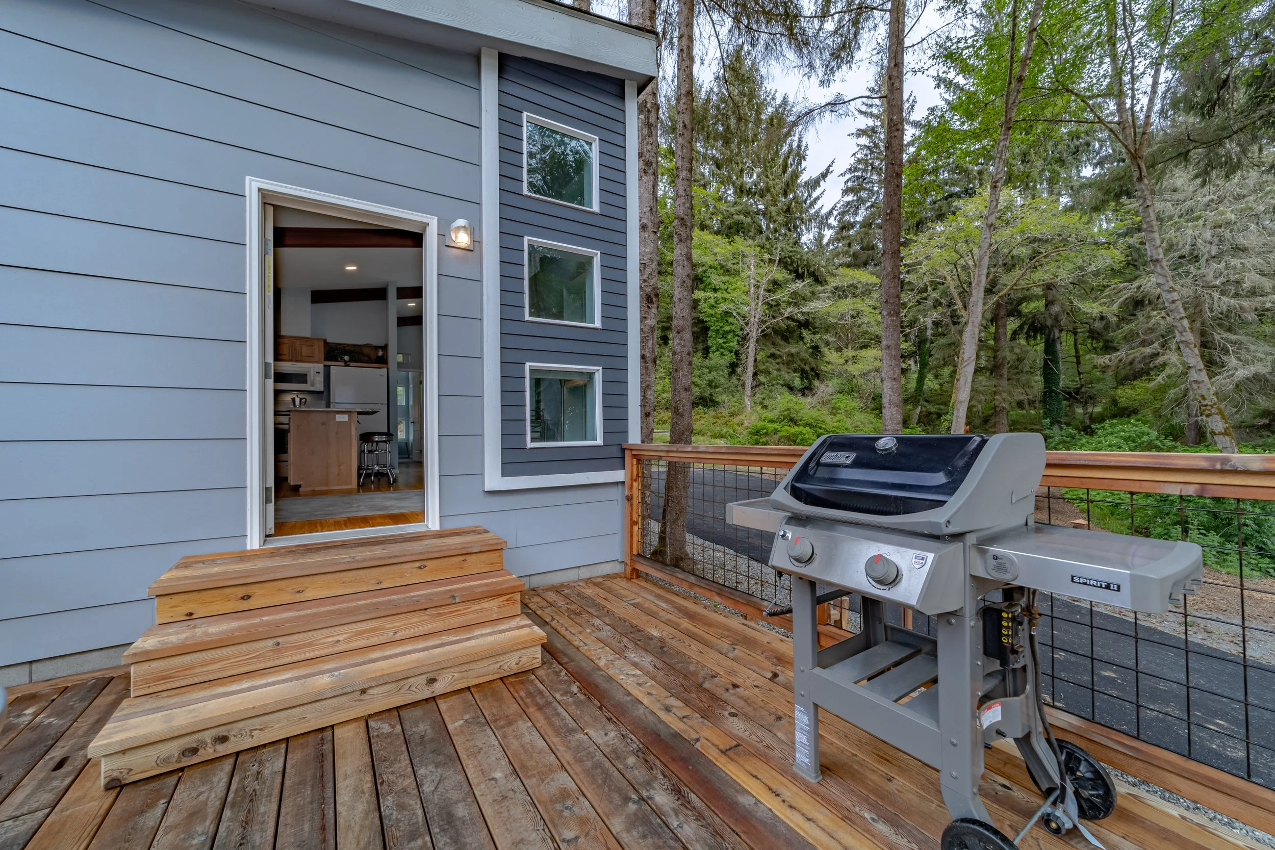 Wooden deck with small steps leading to an open door of a house. Inside the house, kitchen cabinets and a microwave are visible. Next to the door, there are three windows. On the deck, there's a gray propane barbecue grill. The house has light blue h