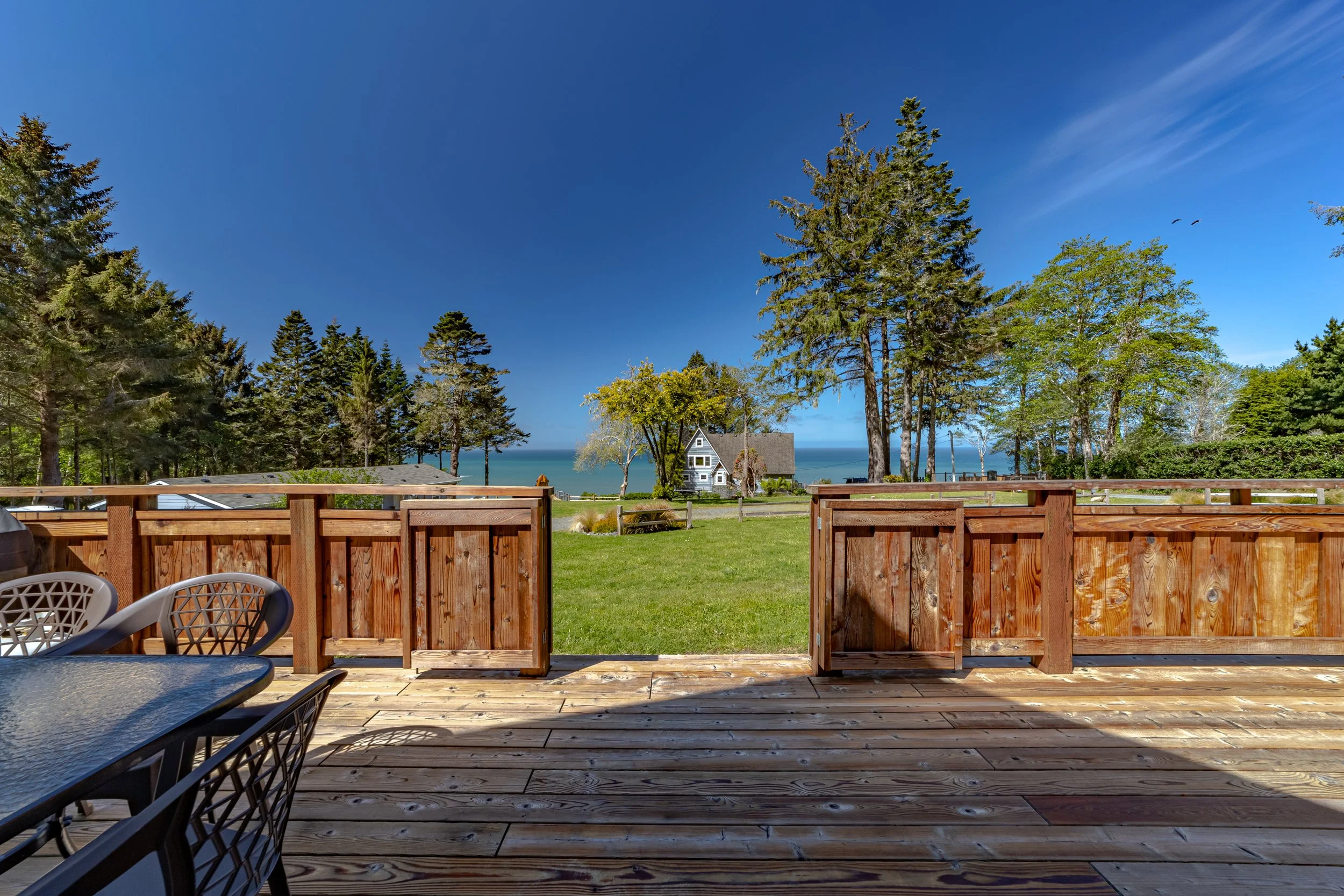 View from a wooden deck looking out over a green lawn with tall trees, a small house, and the ocean beyond under a blue sky.