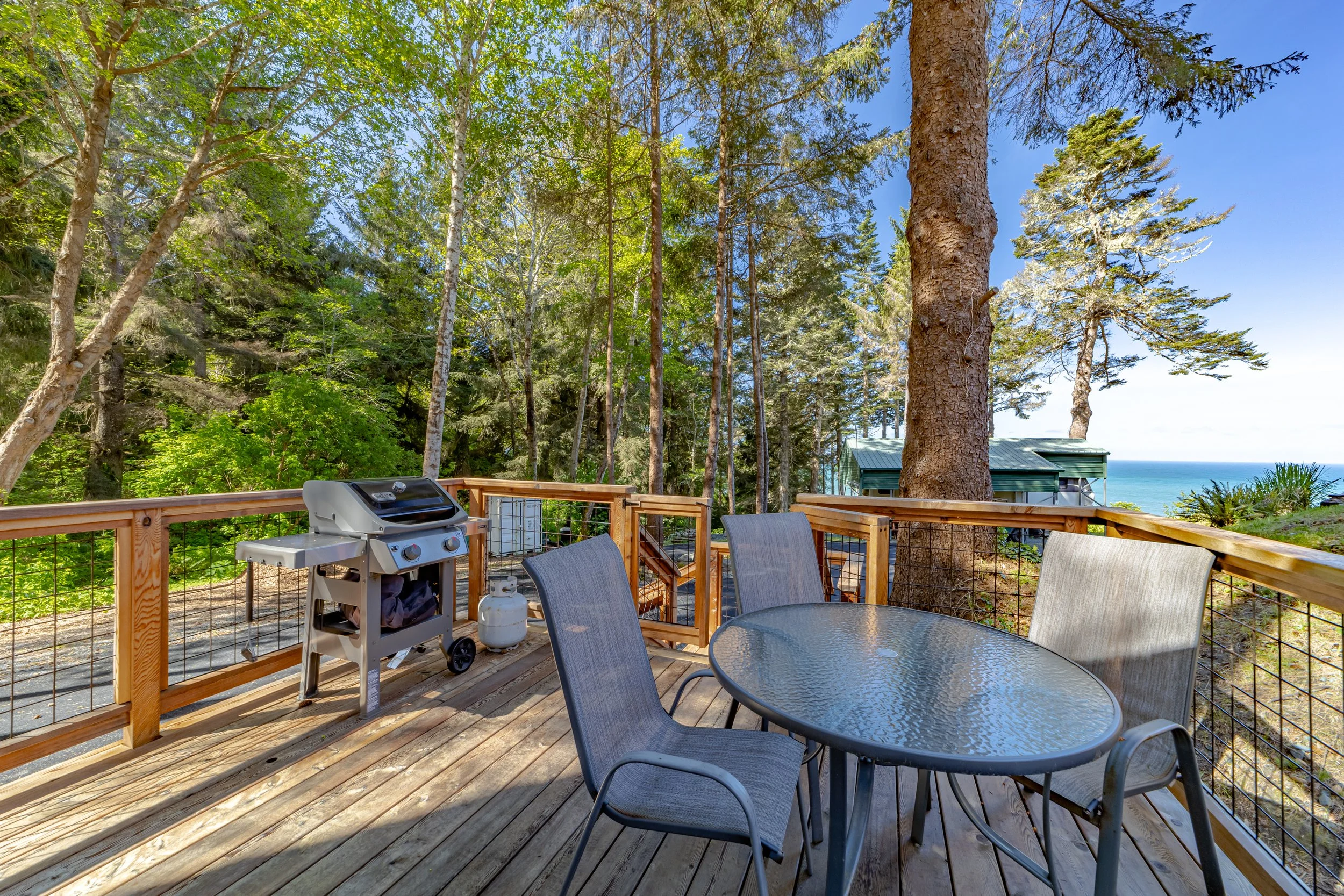 A wooden deck with outdoor furniture and a gas grill, overlooking a forest and the ocean.