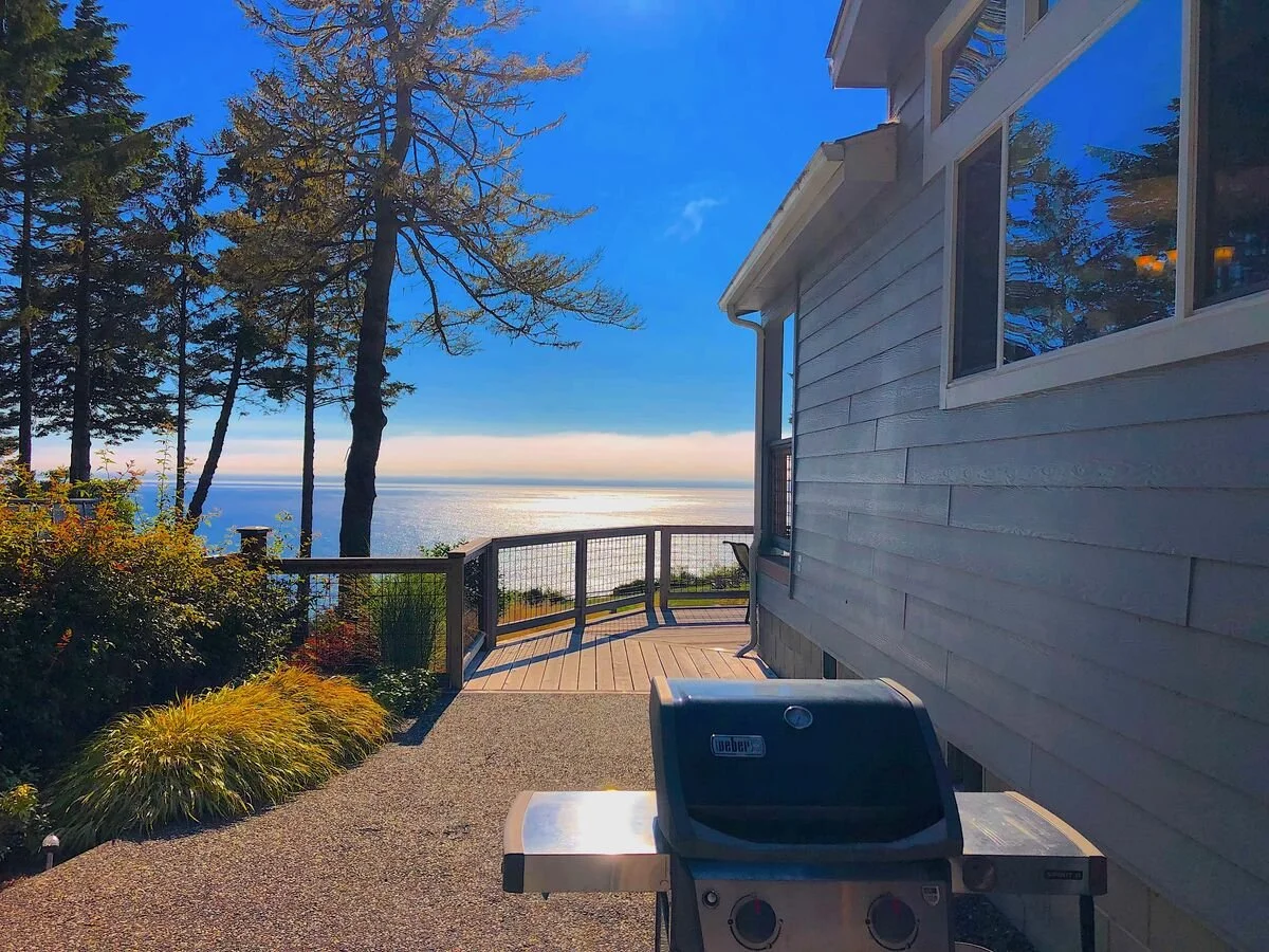 A deck with a barbecue grill overlooking a body of water with trees and a house exterior nearby, under a blue sky with clouds.