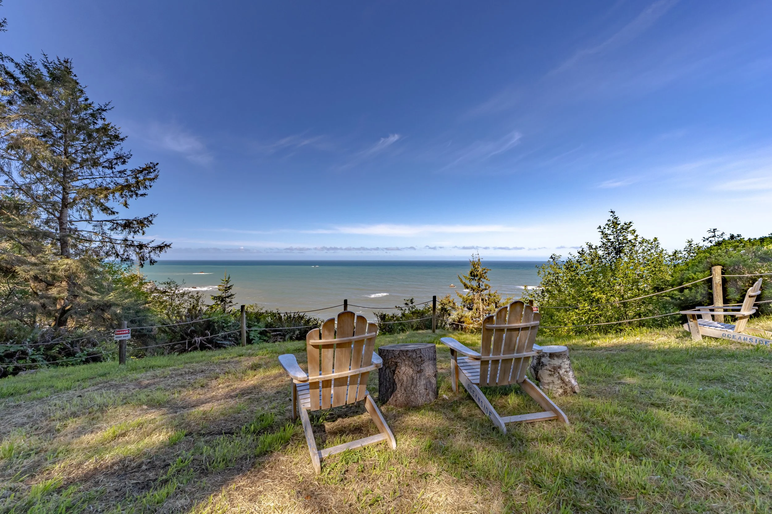Two wooden Adirondack chairs and a tree stump on a grassy hill facing the ocean with trees and a clear blue sky in the background.