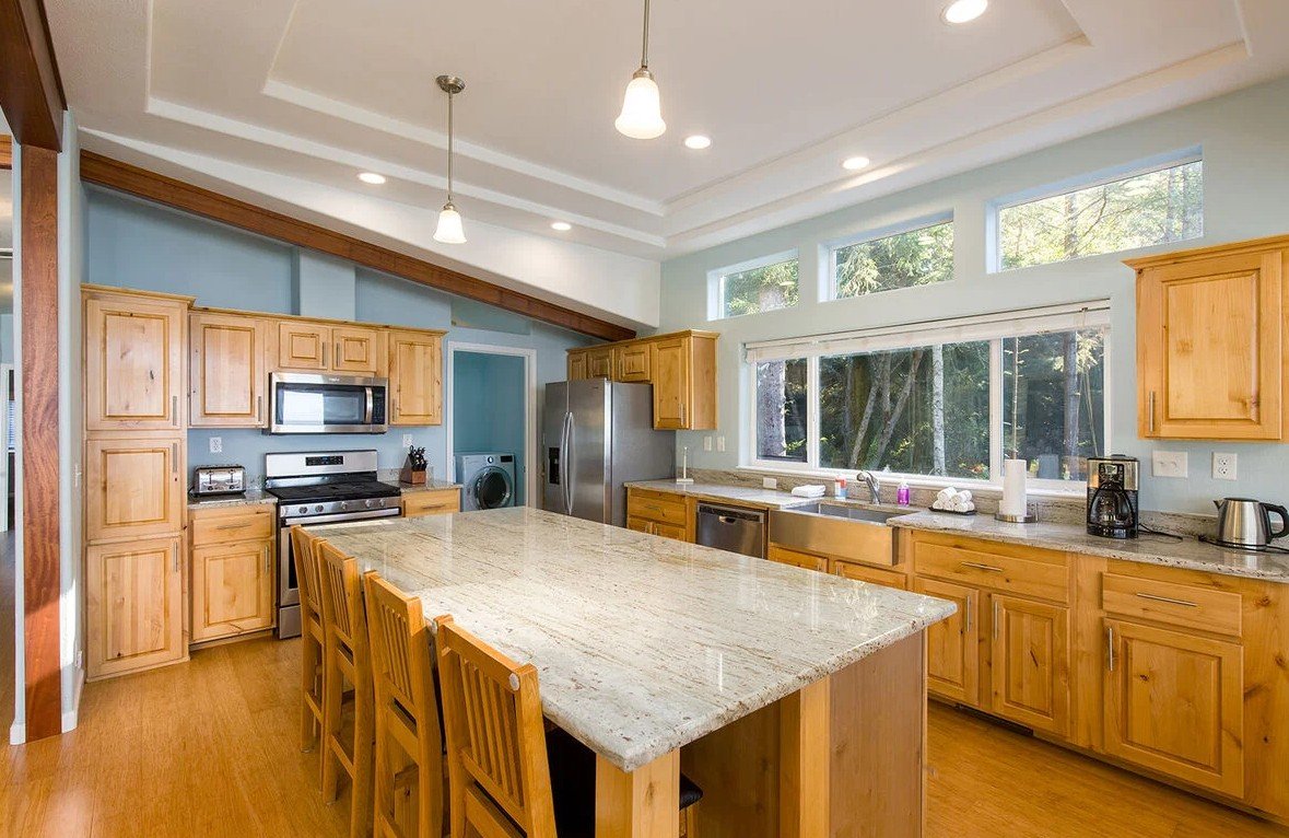 Kitchen with wooden cabinets, a large granite island, stainless steel appliances, and a large window overlooking trees.