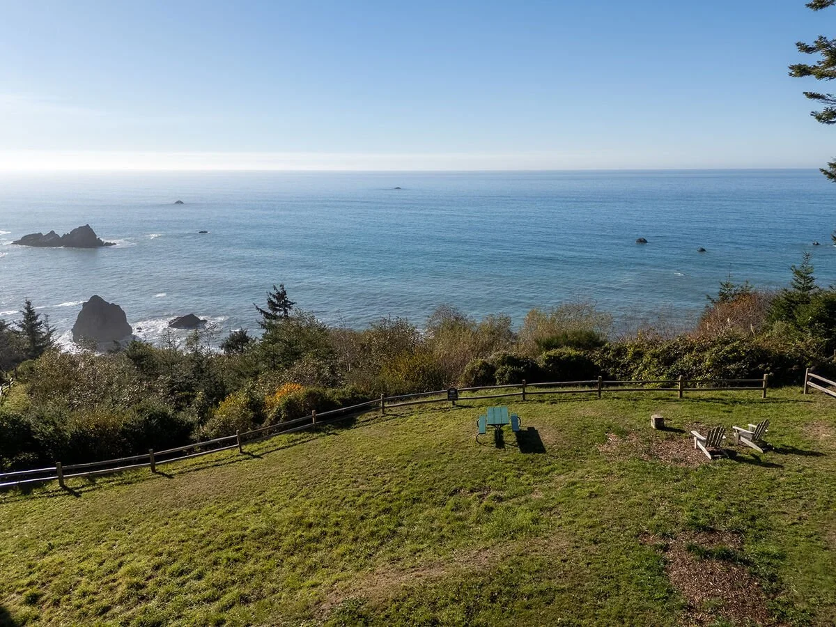 A scenic view of the ocean with rocky formations, seen from a grassy hill with a wooden fence. There are a couple of benches on the hill facing the water.