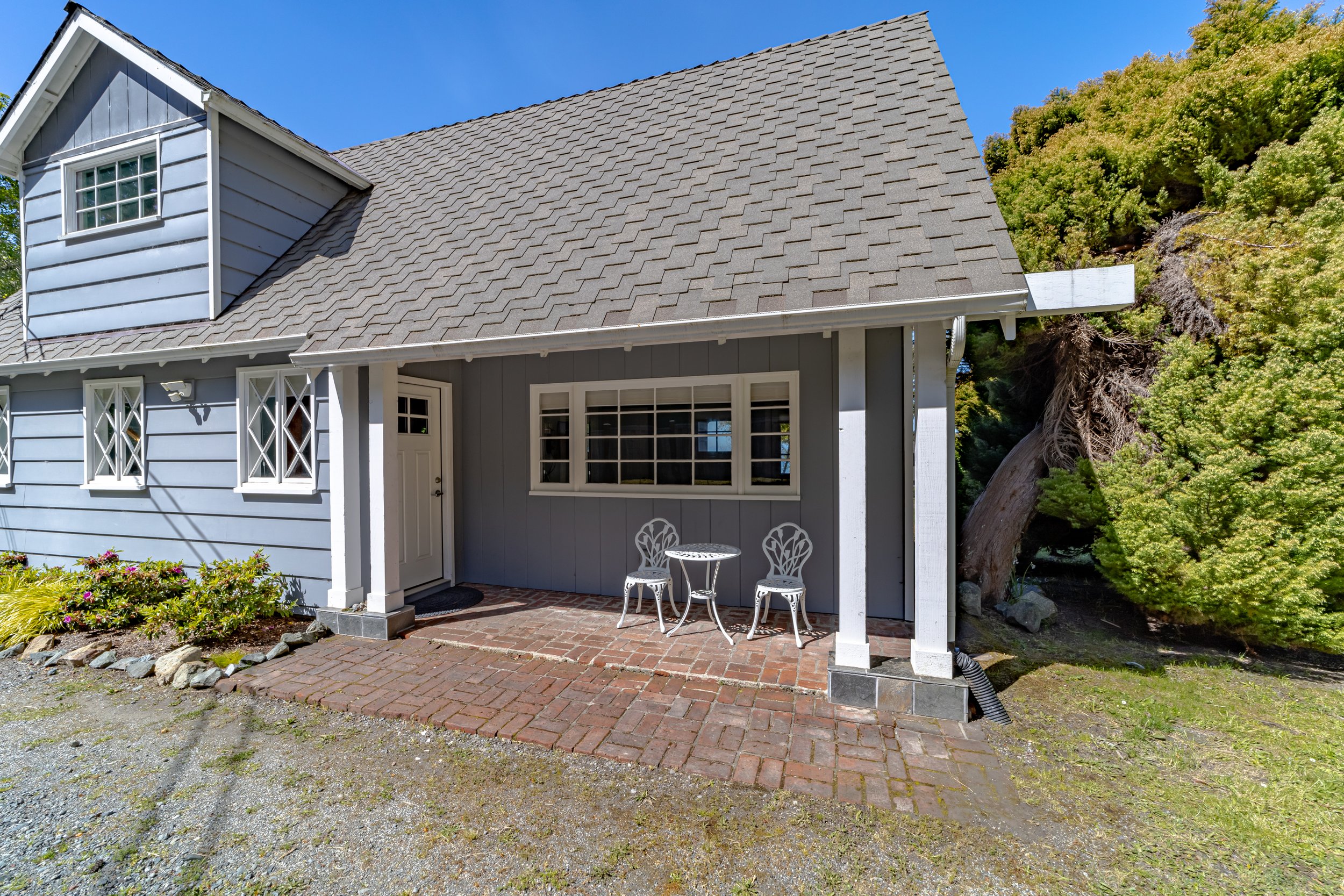 Front porch of a house with a small white metal table and two white metal chairs, a white door, and a large window. The house is painted blue with white trim, and there are green trees in the background.
