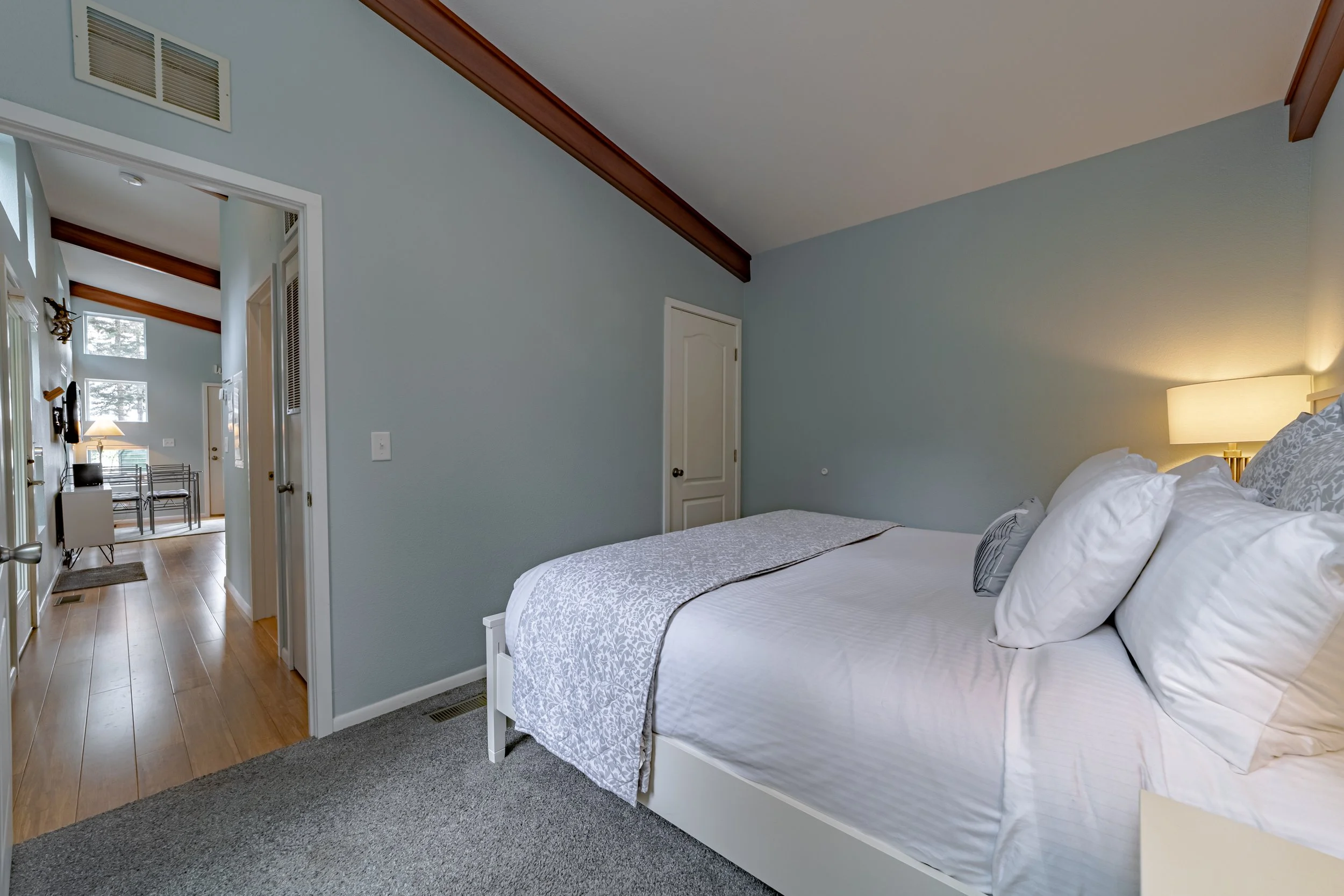 A bedroom with a neatly made bed, white bedding and pillows, a bedside lamp on a nightstand, light blue walls, and a door leading to a hallway with wooden flooring and a sitting area with chairs and a lamp.