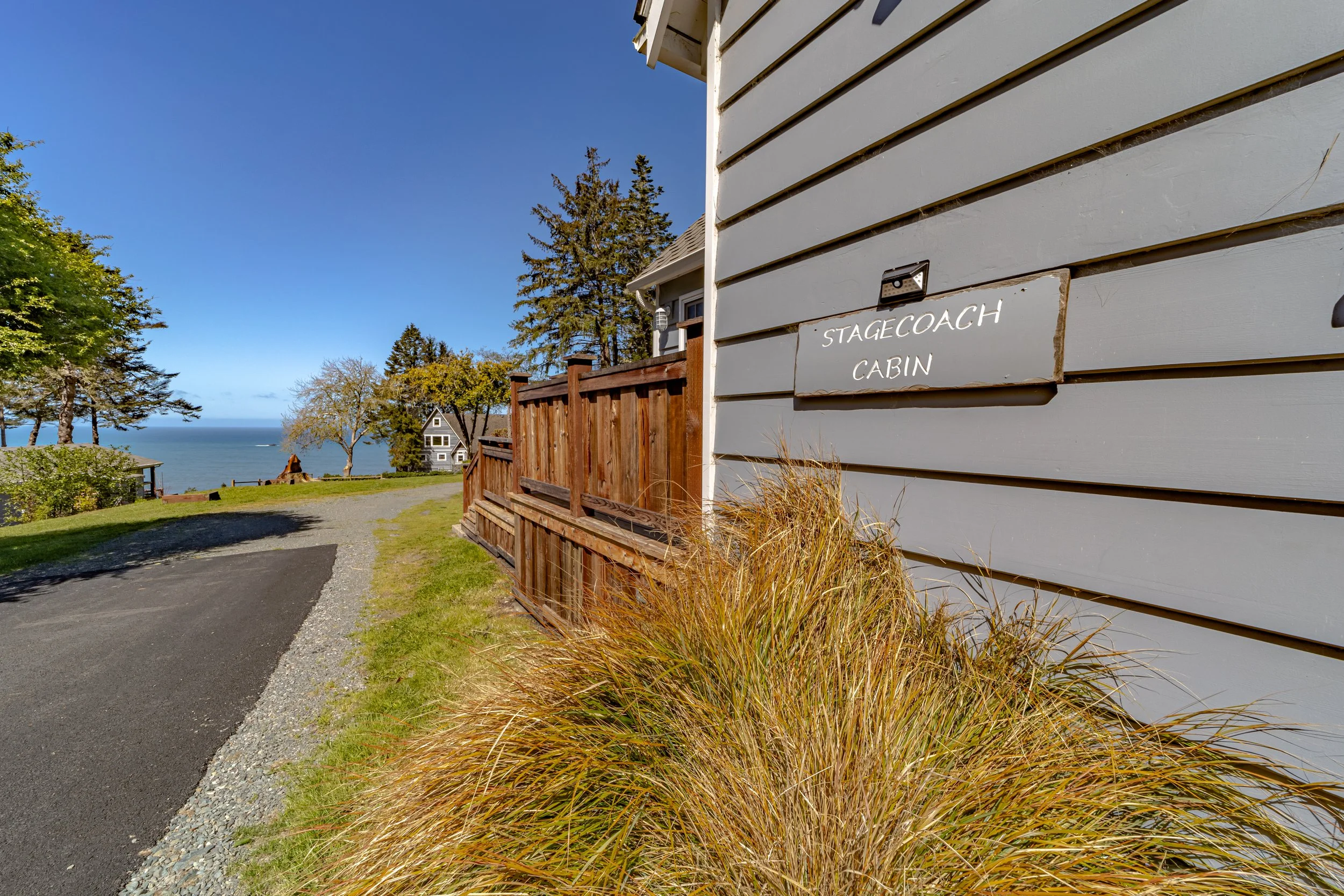 Side view of a house with a sign that reads 'Stagecoach Cabin' on the wall, overlooking a grassy area with trees and a view of the ocean.