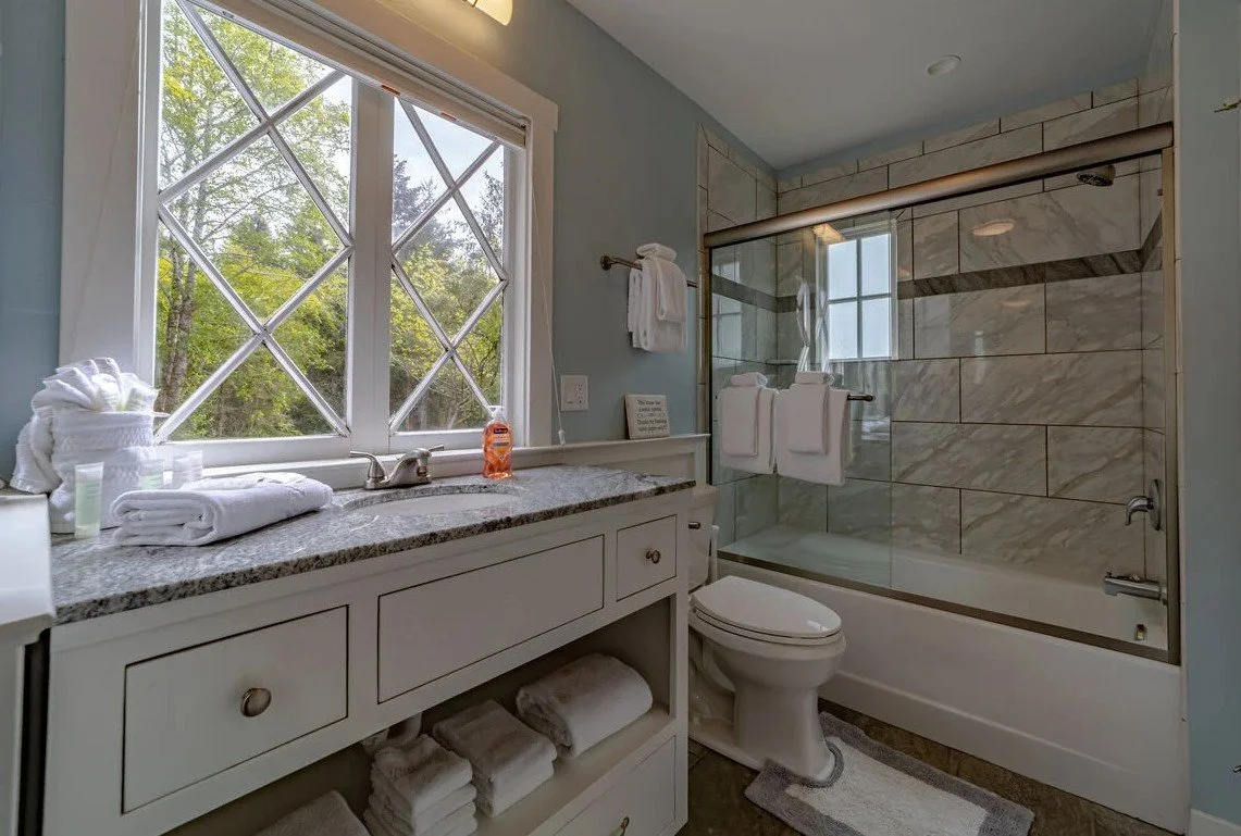 A bathroom with a large window, a countertop with towels, toiletries, and a sink, a toilet, and a glass-enclosed shower with marble tiles.