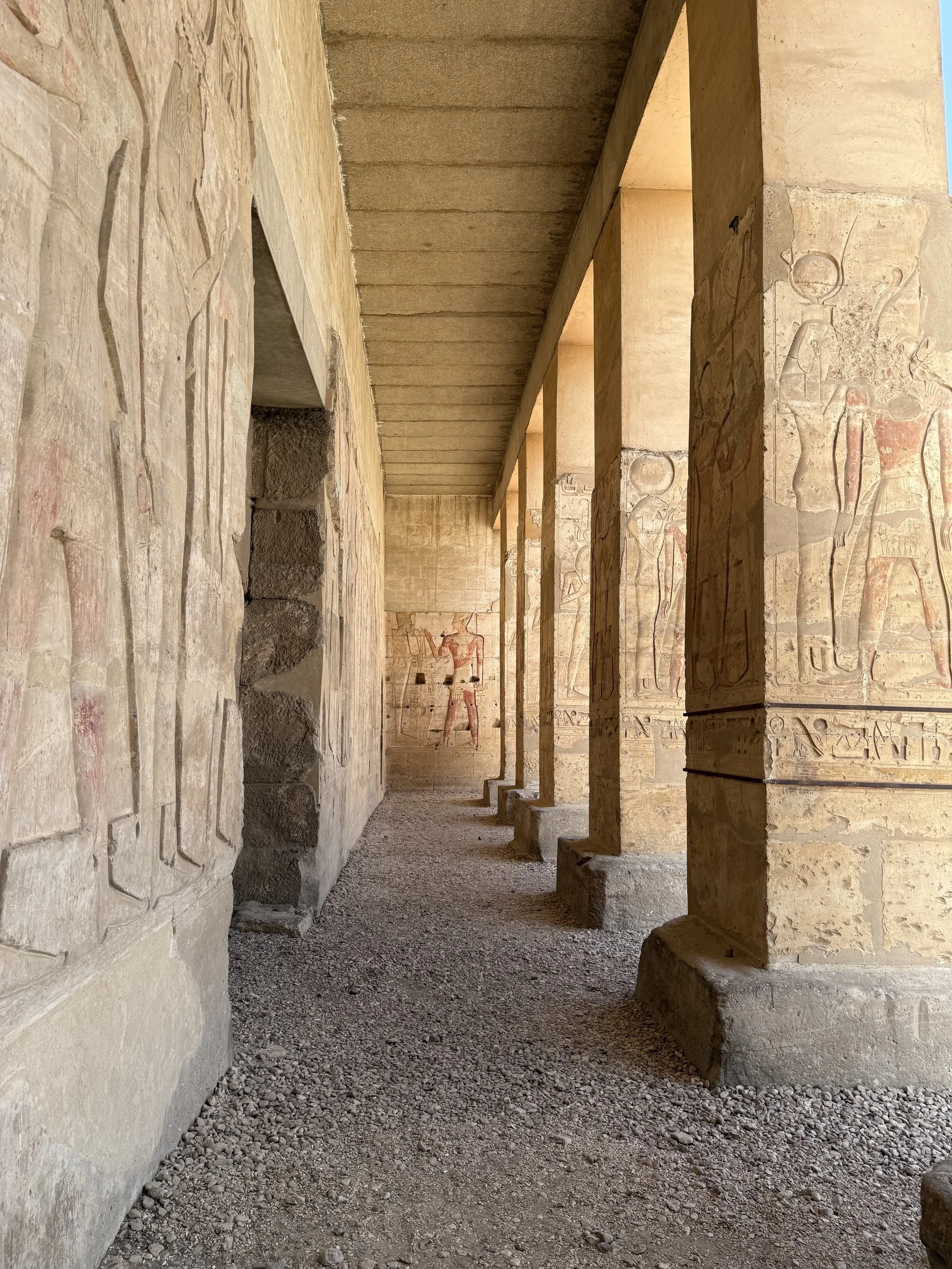 Interior of an ancient Egyptian temple with carved hieroglyphics and paintings on the walls, partially obscured columns, and a gravel floor.
