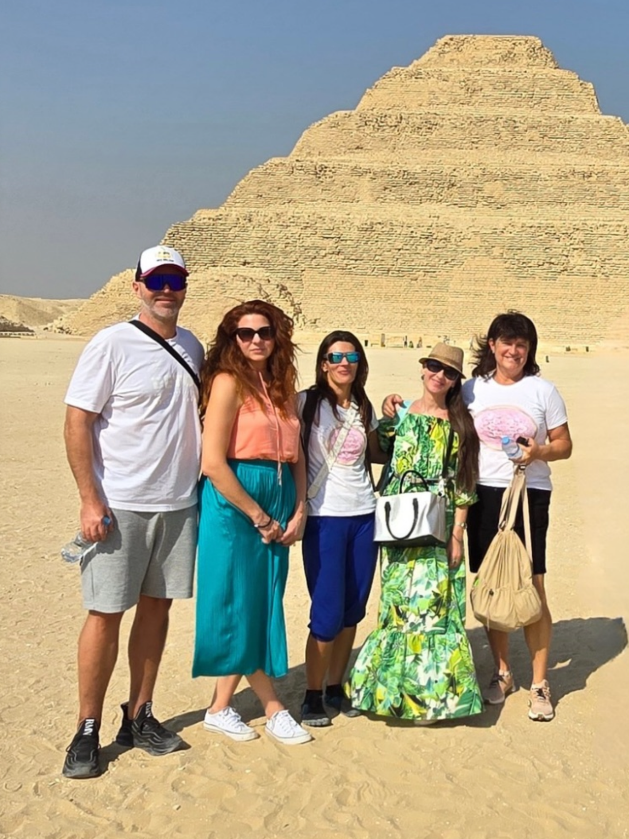 A group of six people posing in front of a pyramid at the desert in Egypt, wearing sunglasses and summer clothes.