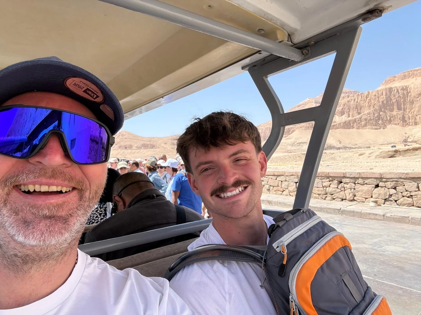 Two men taking a selfie on a golf cart with the Grand Canyon in the background. The man on the left is wearing sunglasses and a cap, partially visible, and the man on the right has a backpack and mustache. There are other visitors and rocky canyon formations under a clear blue sky.