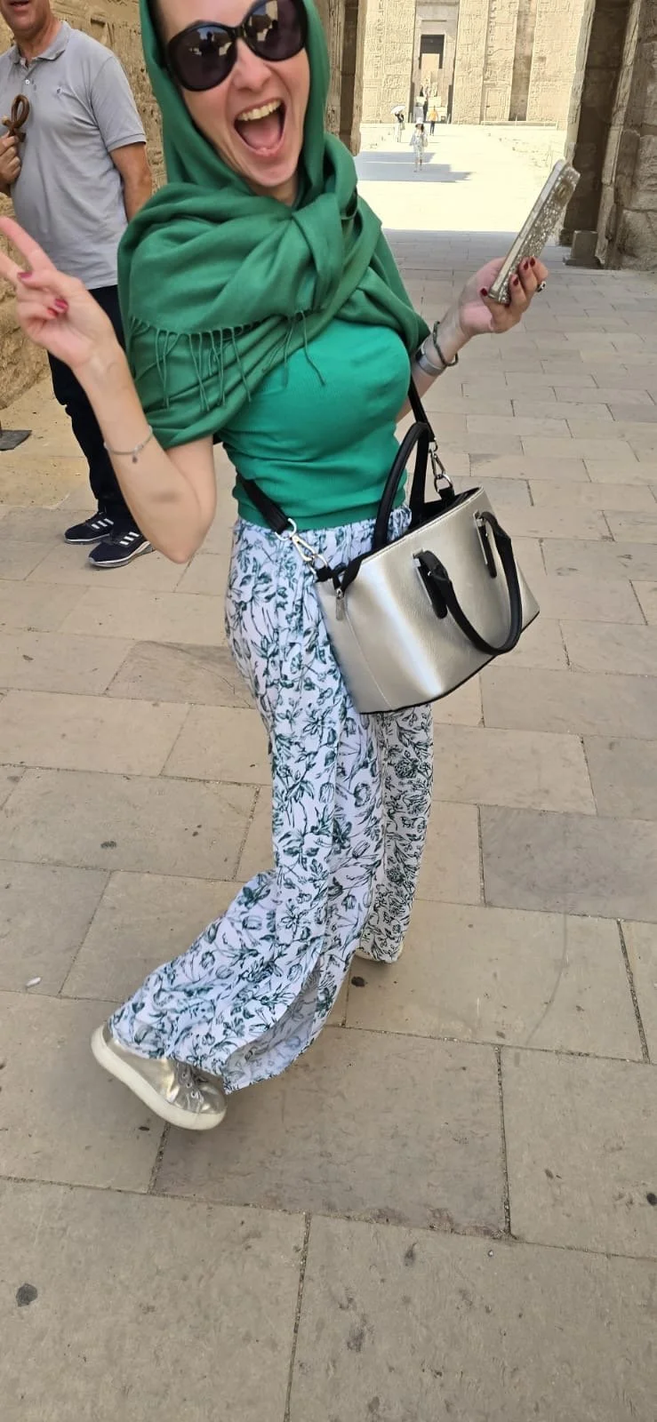 A woman wearing sunglasses, a green headscarf, a green top, and patterned pants, smiling and gesturing with her hands while holding a phone and a metallic purse in a historic stone building or city street.