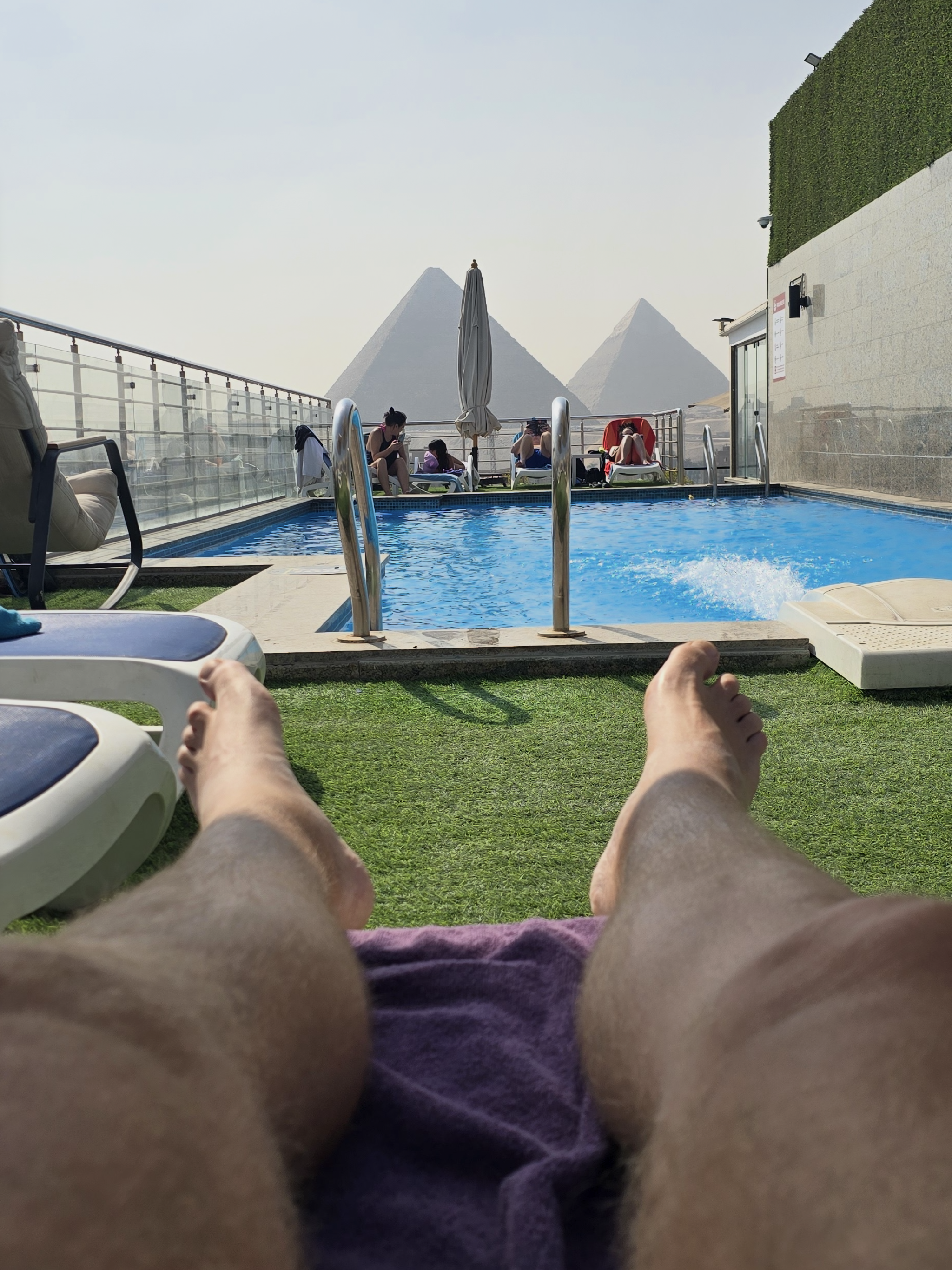 View of a swimming pool area taken from the perspective of someone lying down on a sun lounger, showing their legs and feet in the foreground. In the background, there are people sitting by the pool, some on lounge chairs, with pyramid-shaped structures and a hazy sky in the distance.
