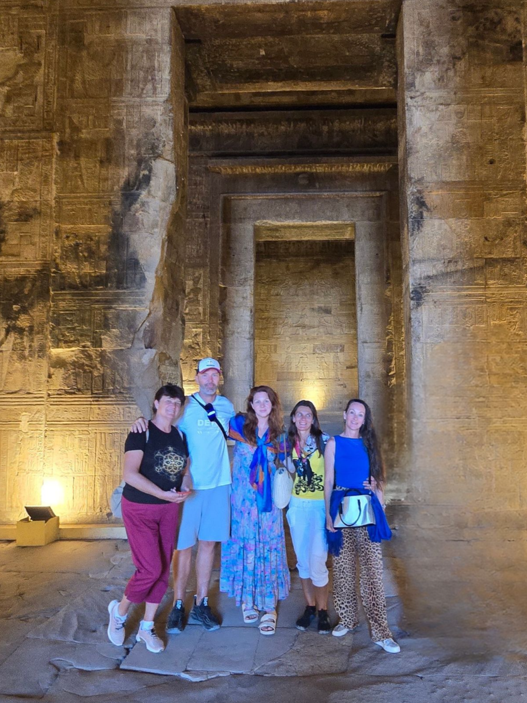 A group of six people standing in front of an ancient Egyptian wall with hieroglyphics, in a dimly lit archaeological site or museum.