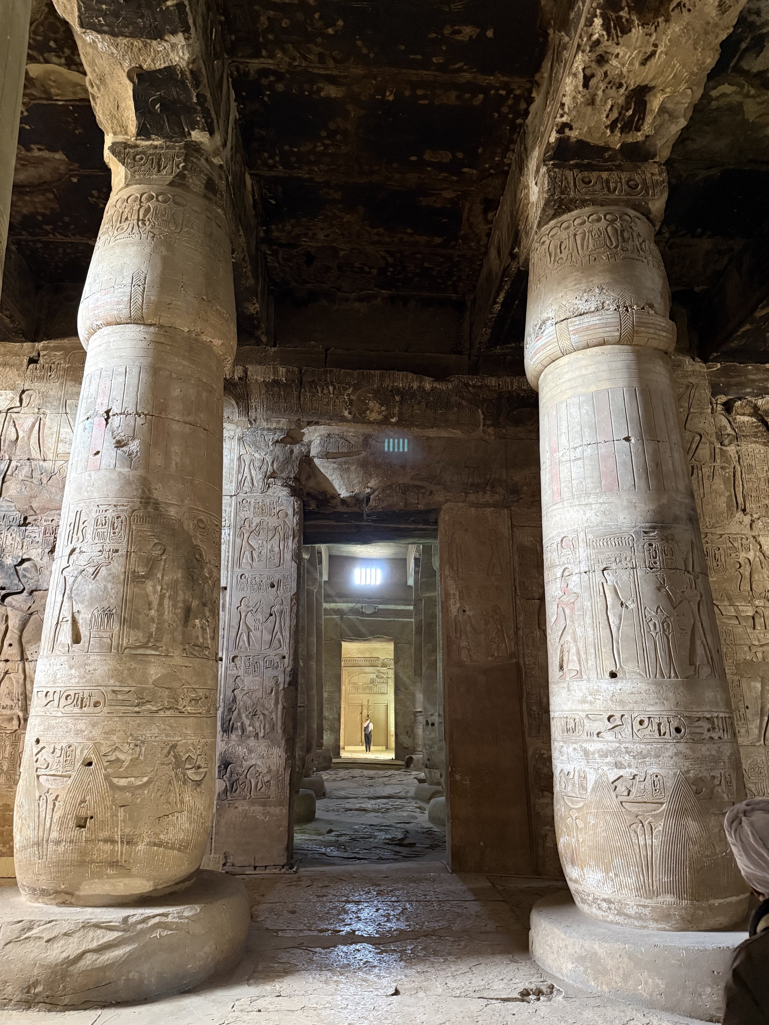 Ancient Egyptian temple interior with massive, hieroglyphically inscribed columns leading to a doorway, with a person standing at the entrance.