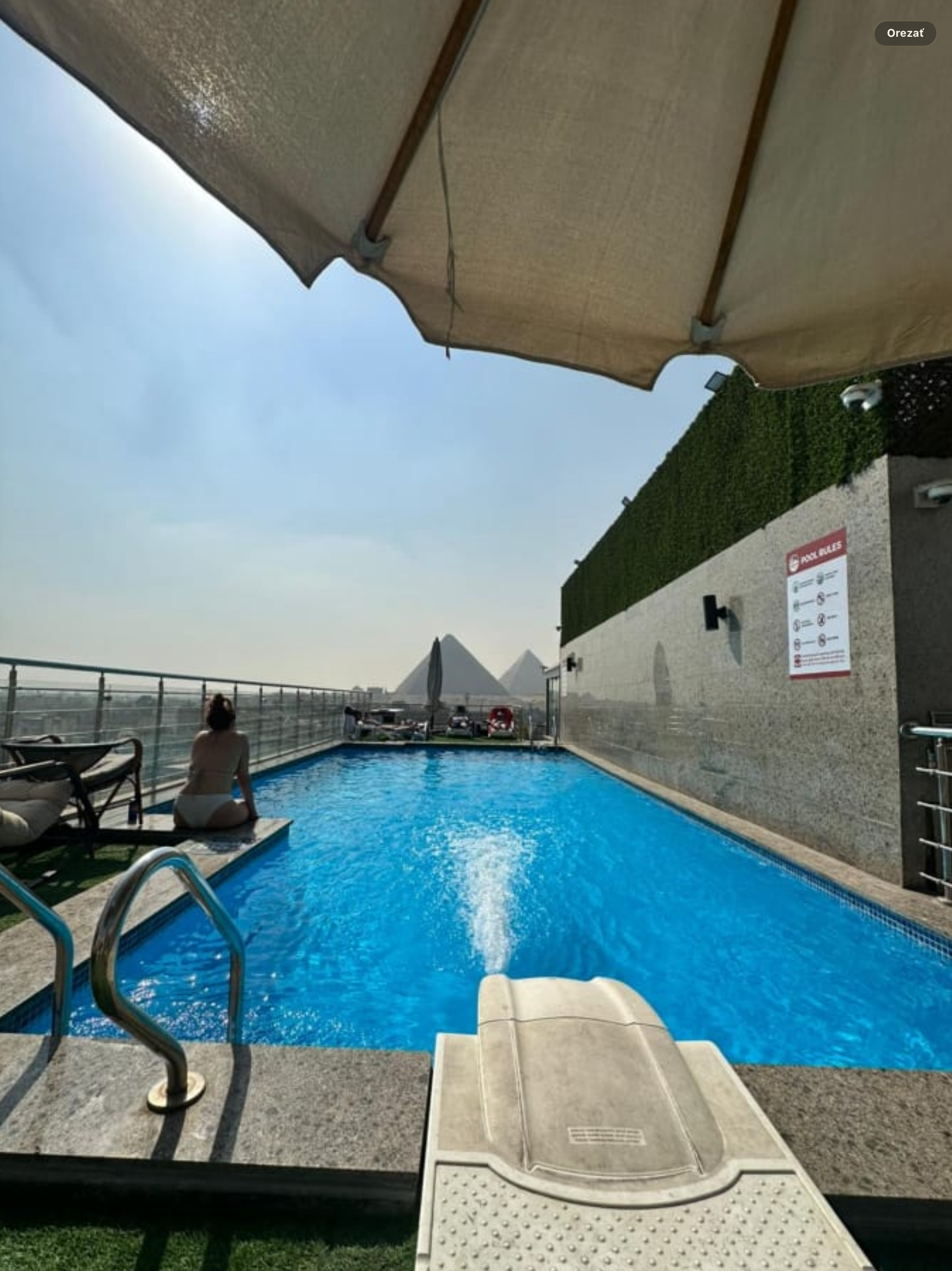 An outdoor swimming pool with a metal handrail and a small beach entry, surrounded by a concrete deck. There are lounge chairs on the deck, a woman sitting near the pool, and a parasol providing shade. The background includes a tall wall with a hedge on top, a sign with pool rules, and a partially visible rooftop area. The sky is clear and sunny.