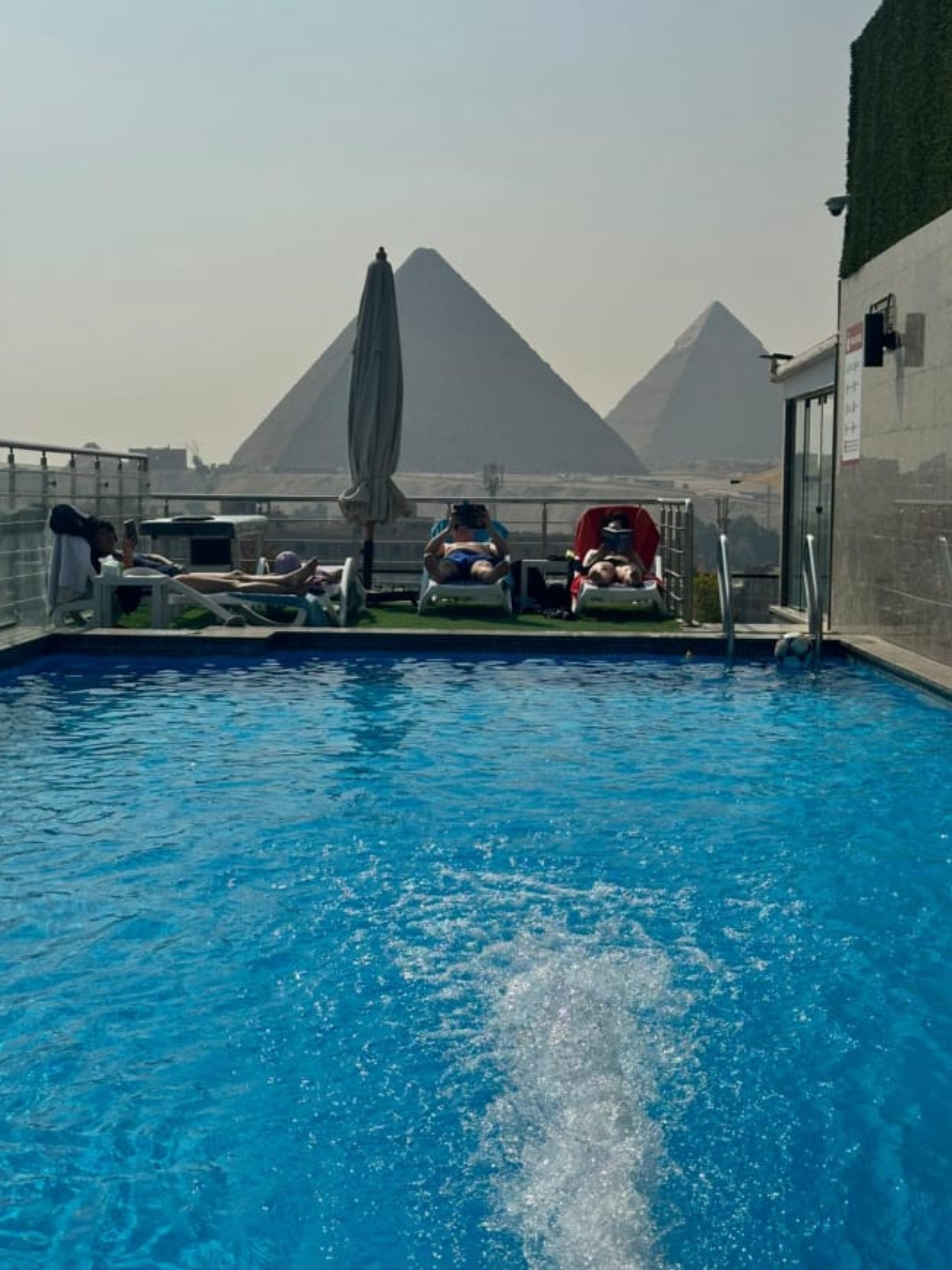 People relaxing by a swimming pool with a view of the pyramids in Egypt in the background.