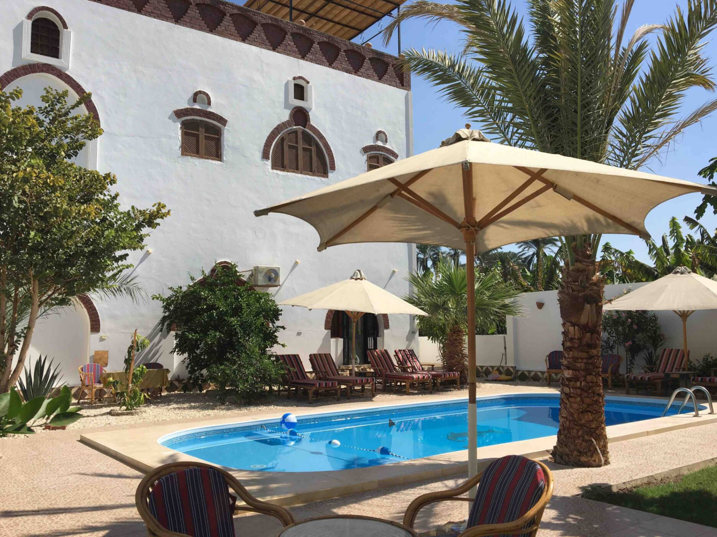 A swimming pool area with a blue pool, multiple lounge chairs with striped cushions, several large umbrellas, lush green trees, and a white building with decorative red brick details, set against a clear blue sky.