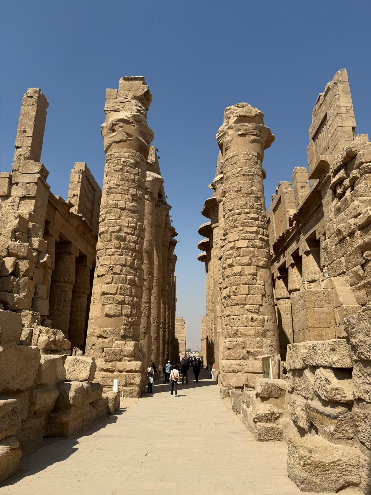 Ancient Egyptian temple ruins with tall stone pillars and hieroglyphic carvings under a clear blue sky, with visitors walking through the site.
