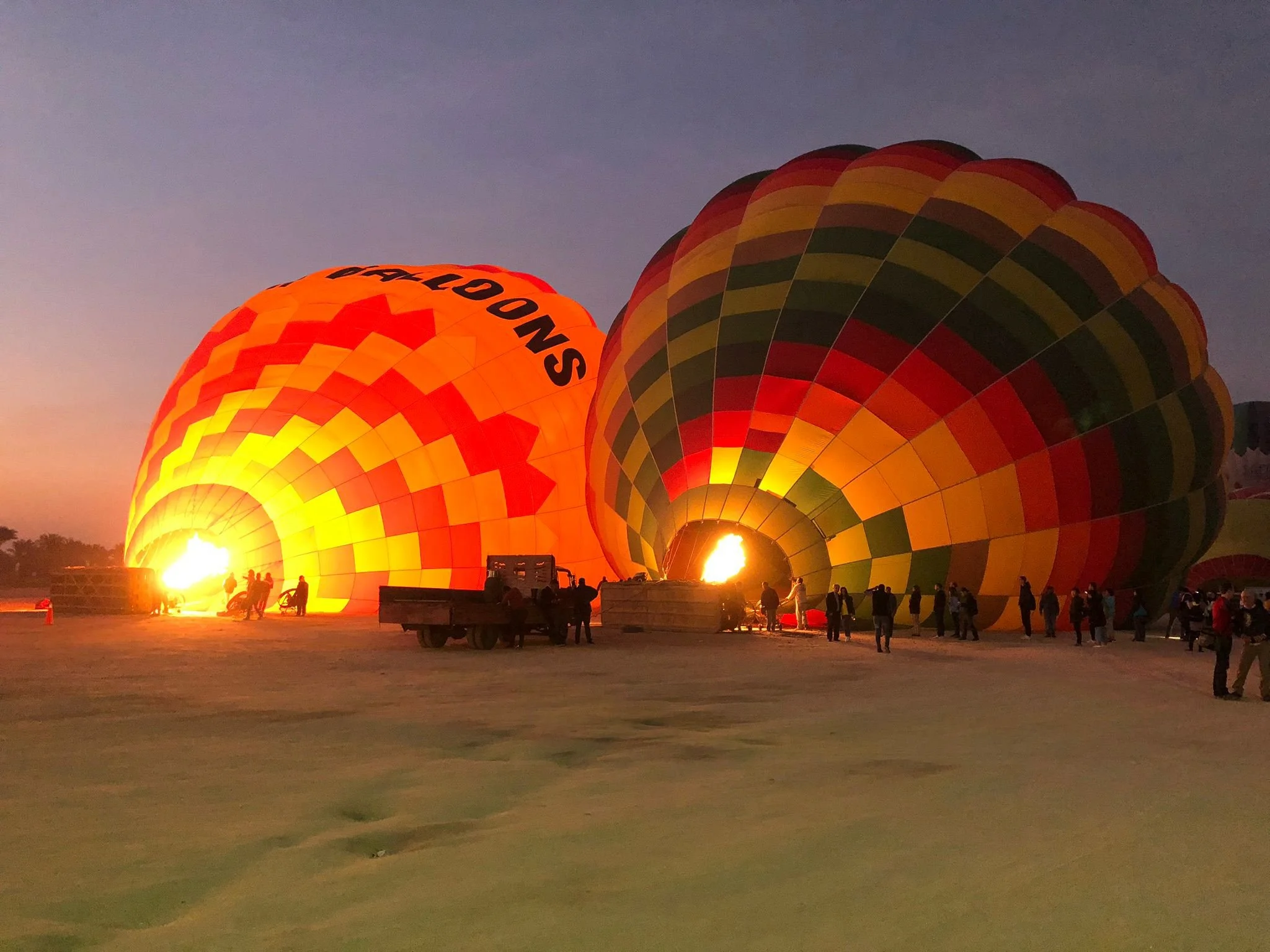 Hot air balloons illuminated from within are being inflated on the ground at dusk or early evening, with people and vehicles nearby.