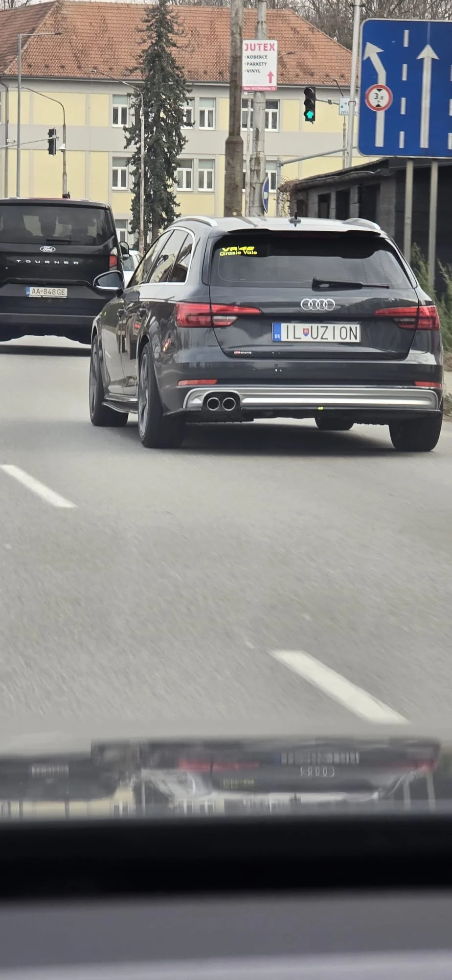A black Audi station wagon with a customized license plate that reads 'ILUZION' driving on a city street.