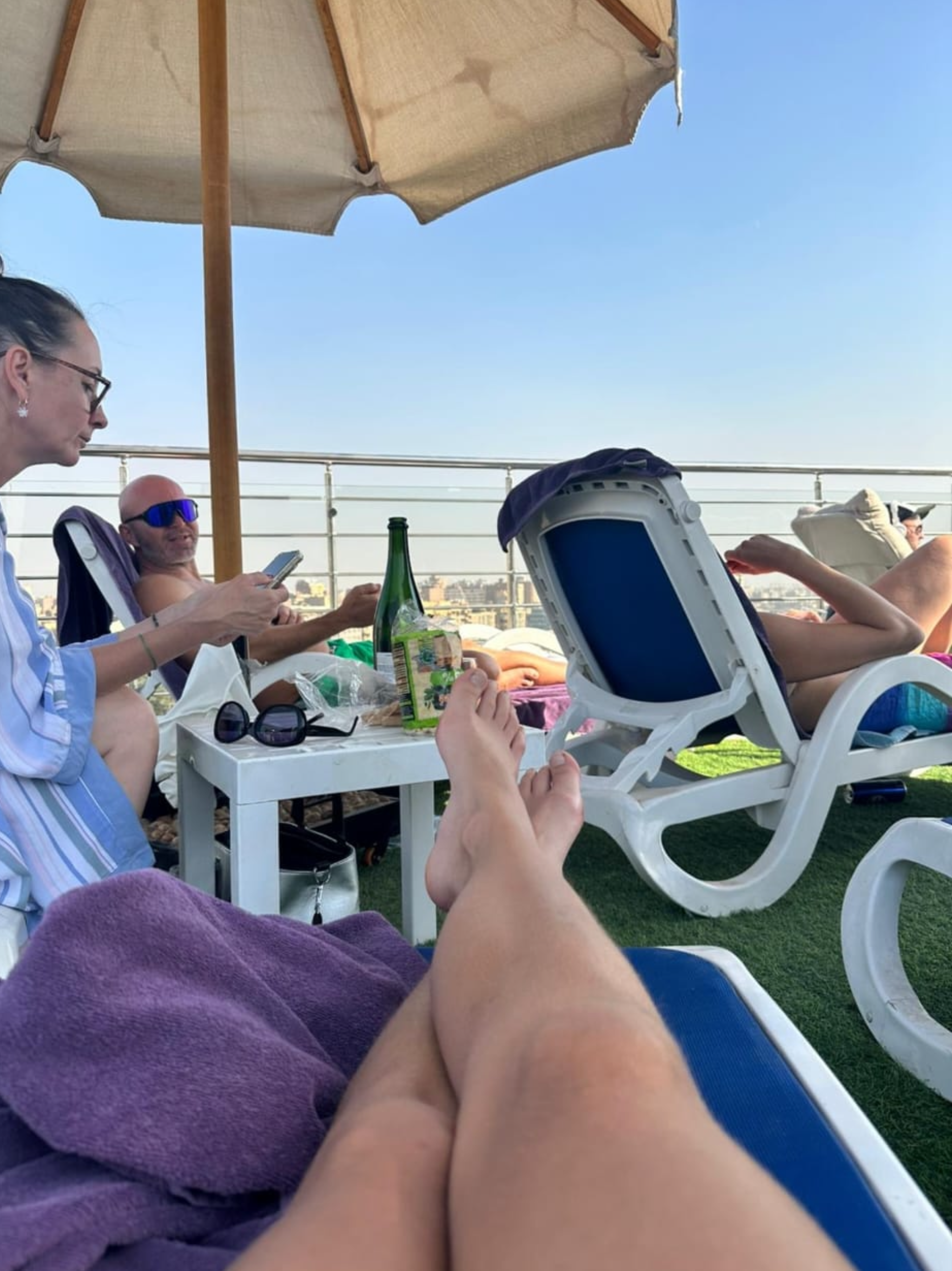 People relaxing on lounge chairs under a large outdoor umbrella on a rooftop with city skyline in the background.