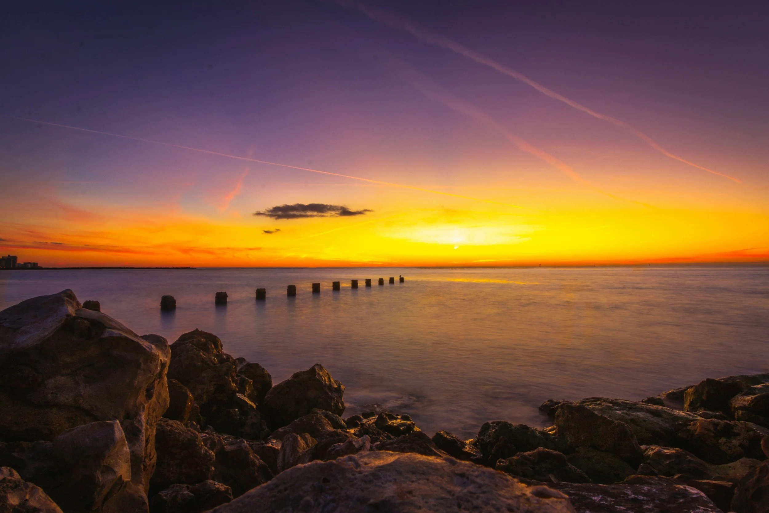 Sunset over water with rocks in the foreground and wooden posts extending into the water.