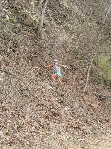Child in a hat and teal clothing sliding down a steep, wooded embankment covered with dry leaves and branches.