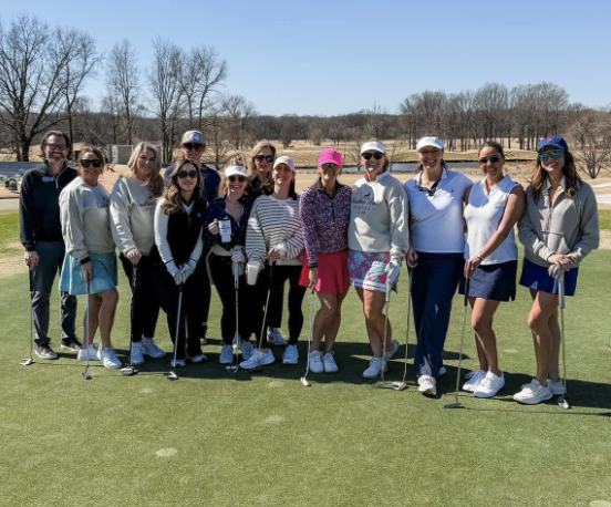 Group of women on a golf course holding golf clubs, dressed in sporty casual attire, with clear blue skies and leafless trees in the background.