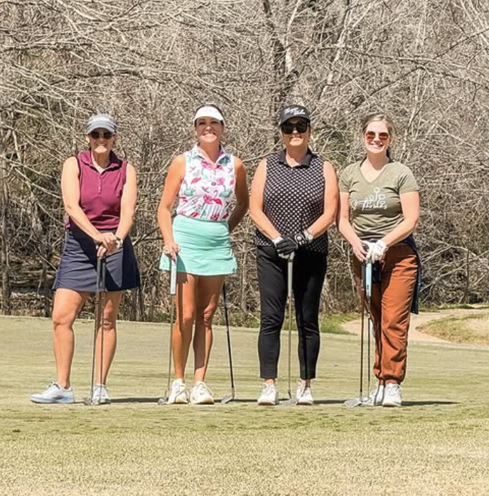 Four women in golf attire standing on a golf course with golf clubs, smiling in front of leafless trees.