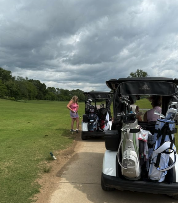 Two golf carts filled with golf bags parked on a golf course path under a cloudy sky, with a woman in a pink top and purple skirt standing nearby.