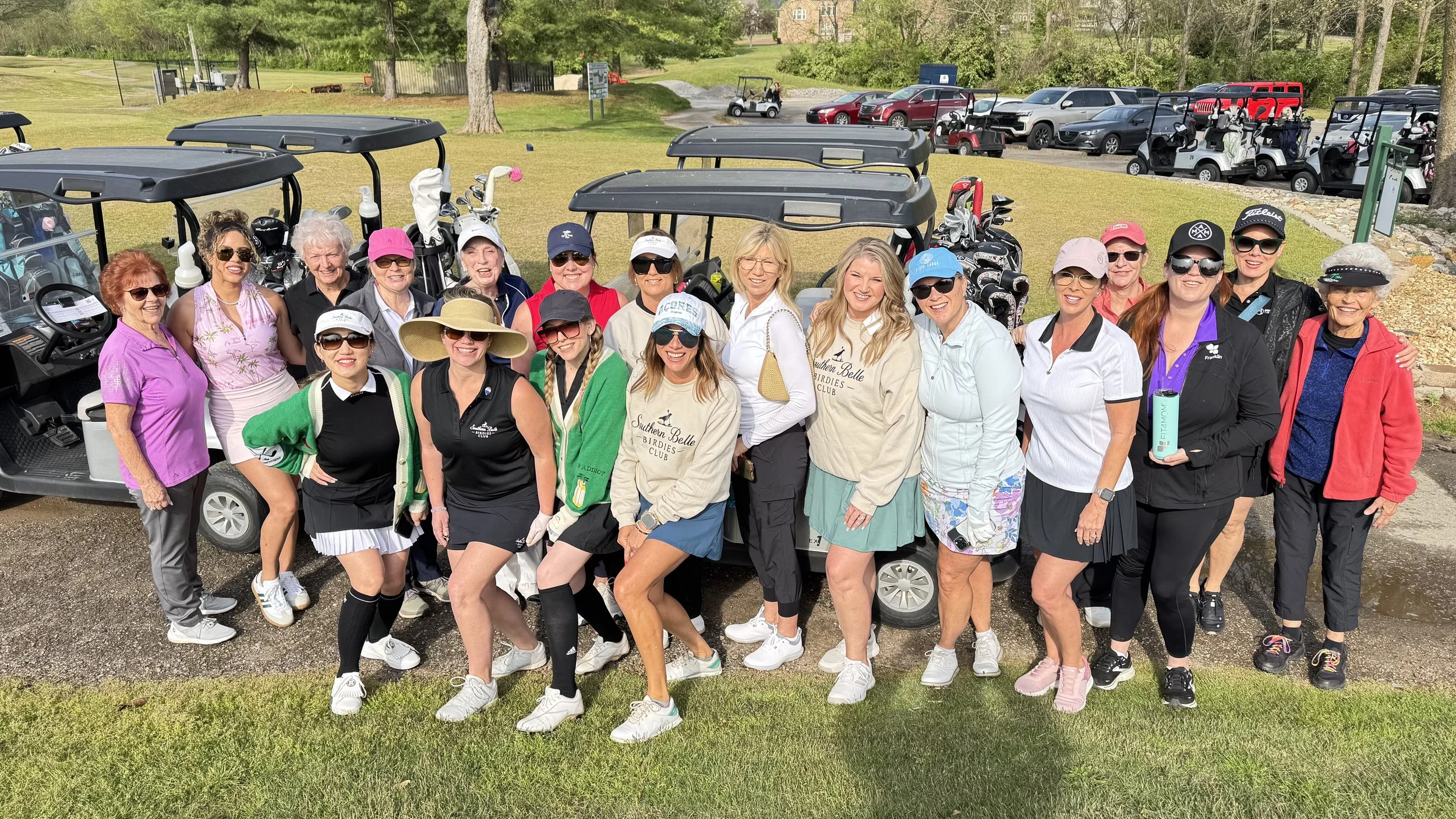A group of women on a golf course, posing in front of golf carts, wearing golf apparel and hats.