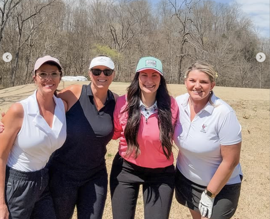 Four women standing outdoors on a golf course, smiling at the camera, dressed in golf attire with hats and gloves.