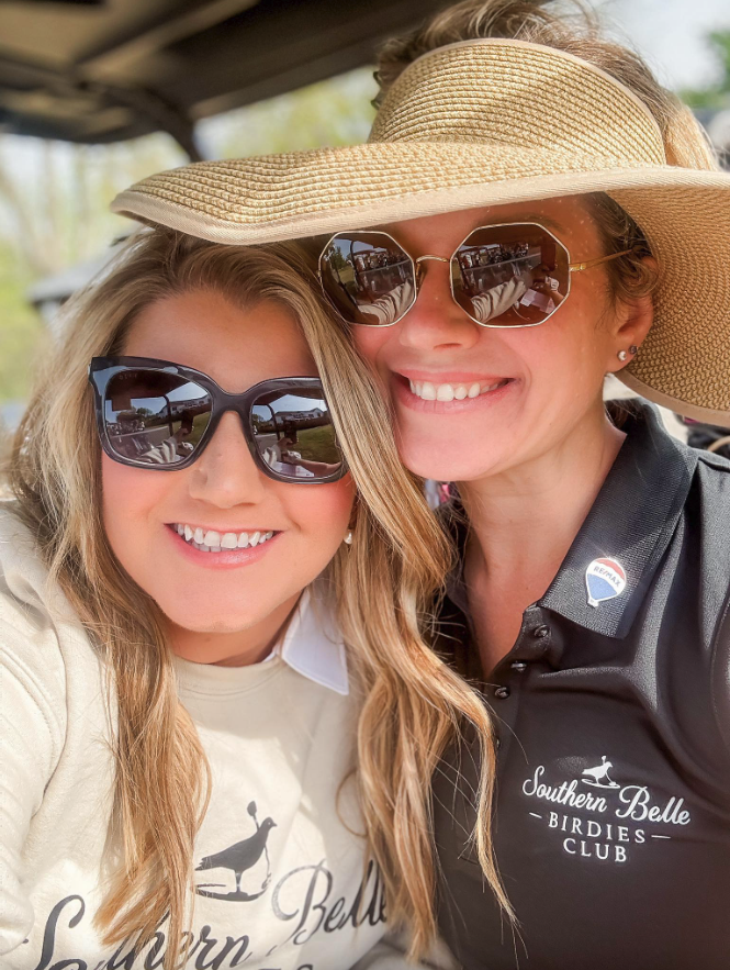 Two smiling women wearing sunglasses and large sun hats, one in a cream shirt and the other in a black polo shirt with Southern Belle Birdies Club logo, taking a selfie outdoors.
