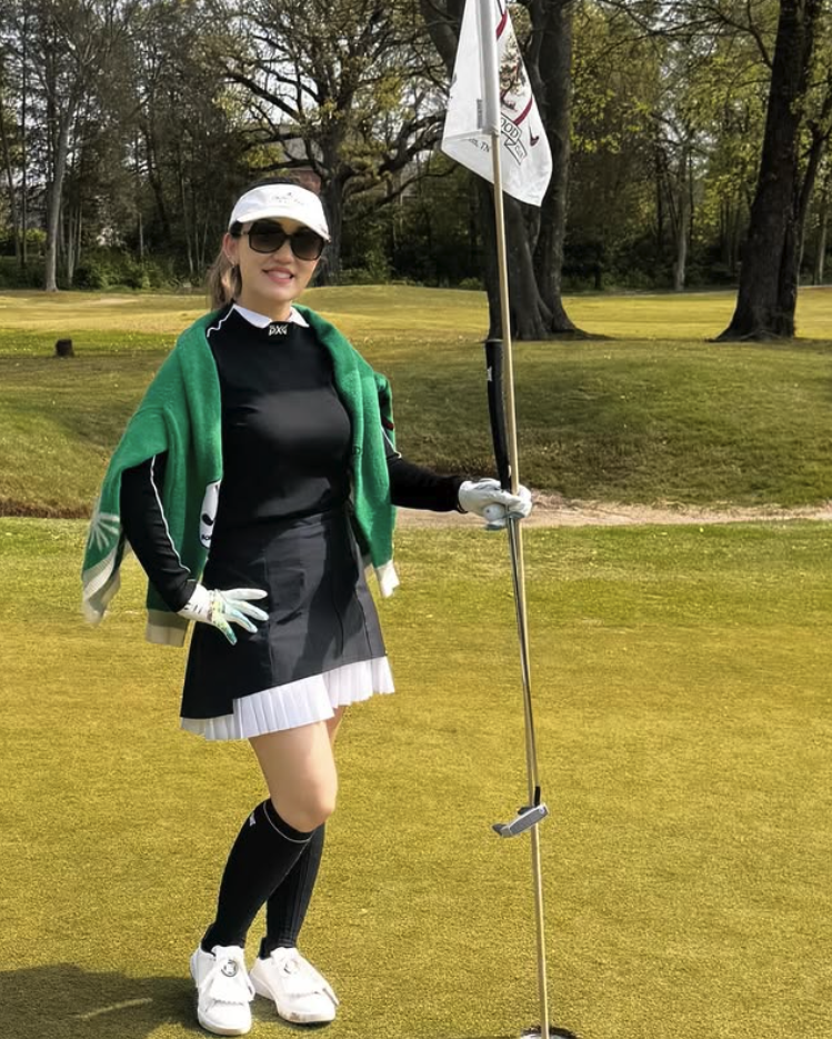 A woman dressed in golf attire standing on a golf course, holding a golf flag. She is wearing sunglasses, a white cap, pedaling clothing, and has a green towel draped over her shoulders.