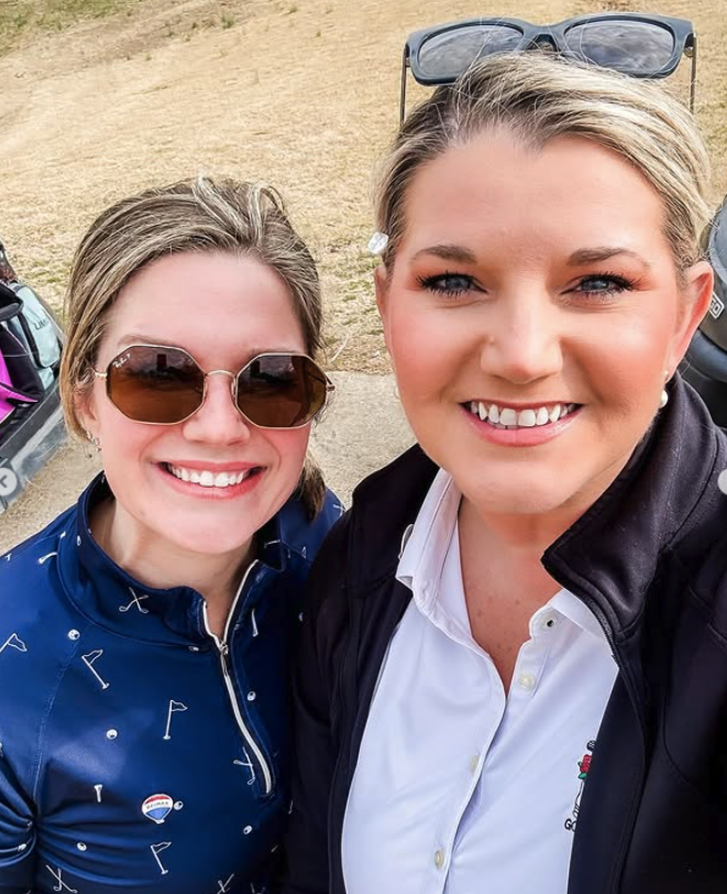 Two smiling women taking a selfie outdoors, one wearing sunglasses and a blue shirt with a golf-themed pattern, and the other wearing a white shirt with a black jacket and sunglasses resting on her head.