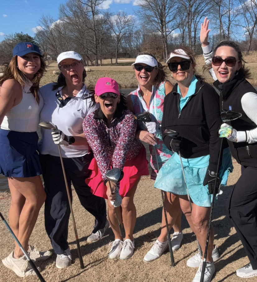 Group of six women smiling and posing on a golf course, dressed in golf attire, holding golf clubs, with trees and a blue sky in the background.