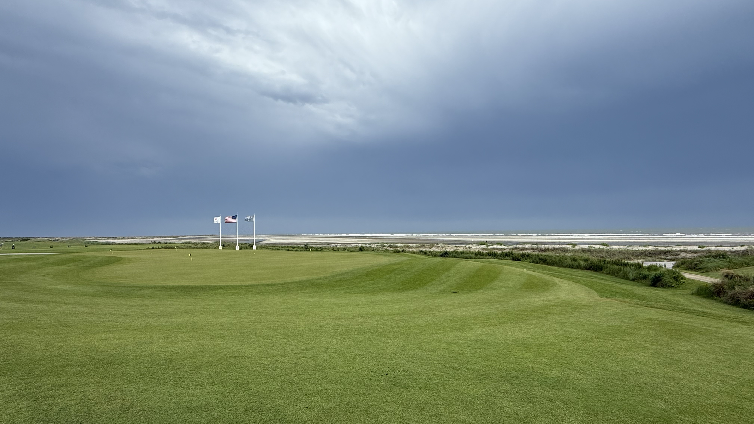 A golf course under a dark, cloudy sky with three American flags flying in the distance.