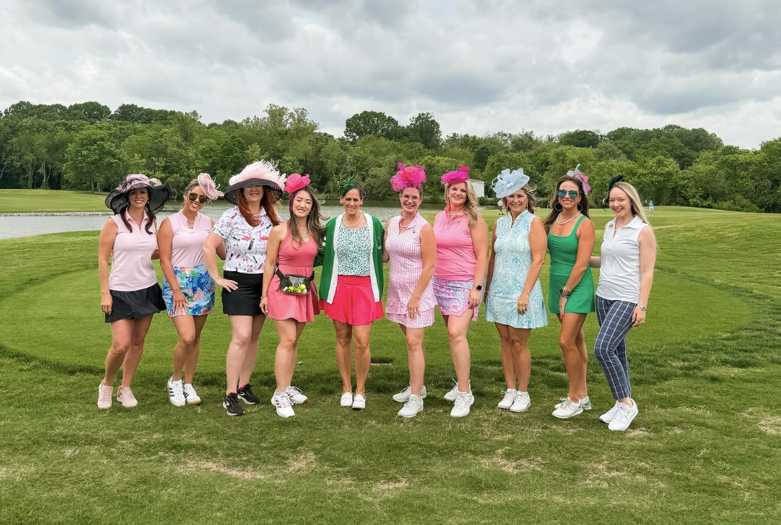 A group of women dressed in colorful, floral, and pastel outfits with wide-brimmed hats and sunglasses, standing together on a golf course with trees and water in the background.
