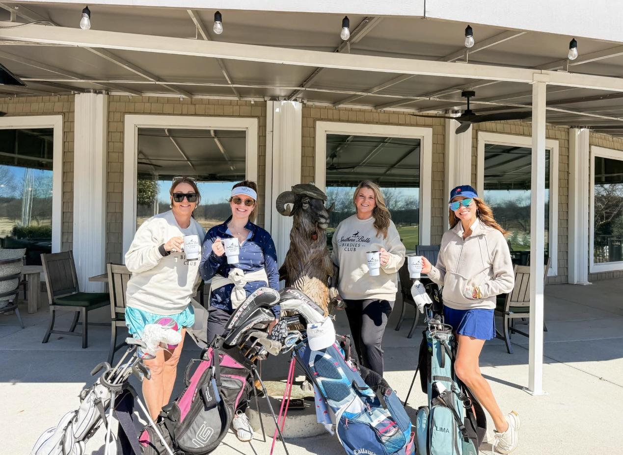 Four women standing with golf clubs and golf bags, holding coffee mugs, posing next to a goat statue outside a clubhouse on a sunny day.