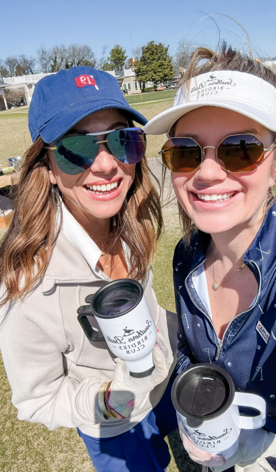 Two women smiling outdoors, wearing sunglasses and caps, holding travel mugs, at a sunny event on a grassy field.