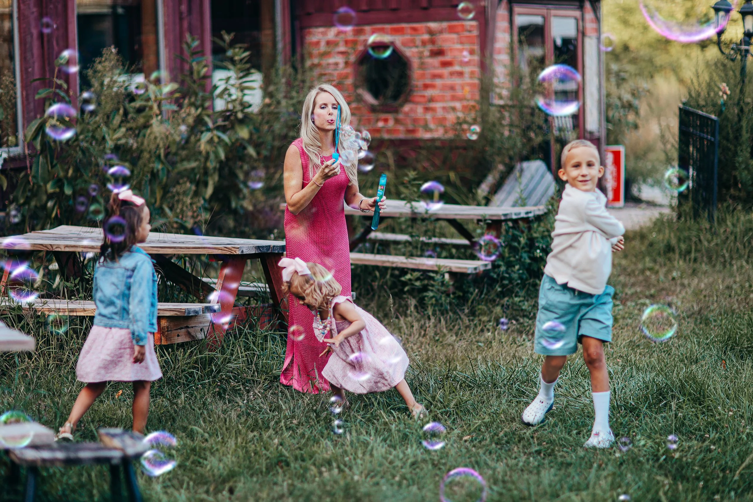 A woman in a pink dress blowing bubbles with children playing outside on a grassy area near a wooden picnic table, with a brick house in the background.