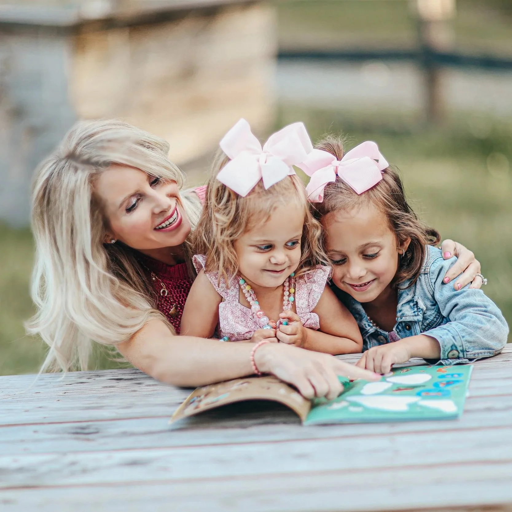 A woman with long blonde hair and a red sweater hugging two young girls with bows in their hair, looking at a colorful book on a wooden table outdoors.