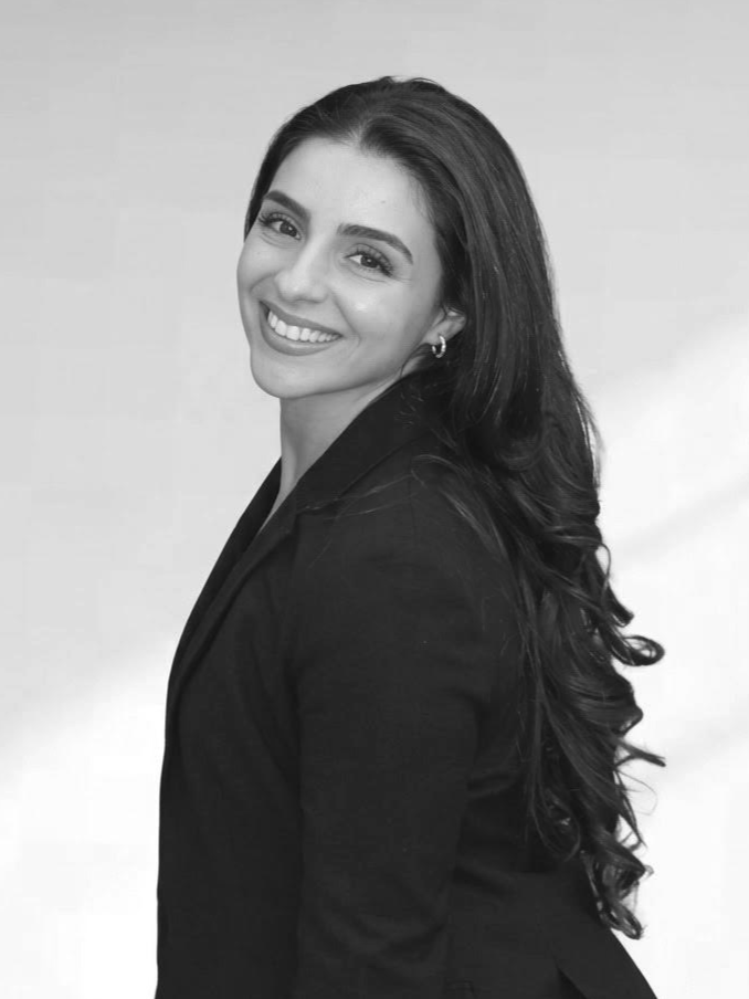 Black and white portrait of a smiling woman with long wavy hair, wearing a blazer, looking over her shoulder.